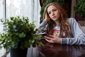 Girl sitting at a table and holding phone while looking sadly out the window.