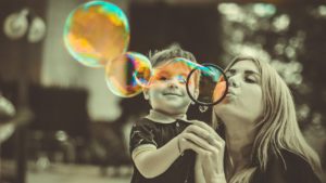 Young boy holding a bubble wand while his mother blows bubbles.