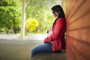 Lone pregnant woman in red jacket sitting on a bench, leaning against a brick wall