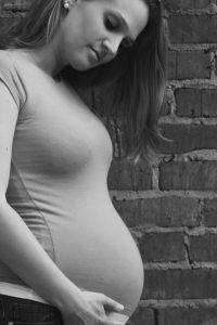 Black and white photo of a young pregnant girl leaning against a brick wall and gently holding her stomach.