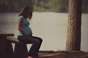 Pregnant woman sitting on a park bench in front of a body of water, contemplating open adoption.