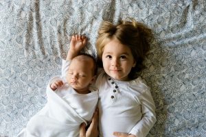adopting a second child. A young boy laying on a bed with his arm around his infant brother. Both are wearing white.