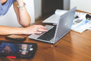 close up of a woman's arms typing on a laptop, searching "How to Find Adoptive Parents." A tablet and some magazines are also on the table.