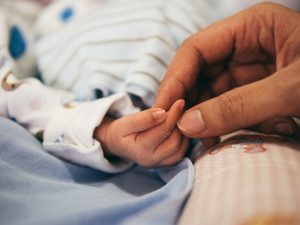 Become Adoptive Parents. Closeup of a mother's hand holding her baby's hand. There is a white blanket with thin stripes covering the child. The baby is wearing a long sleeve white onesie.