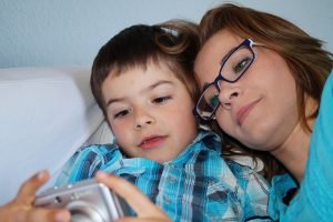 Bonding With Your Adopted Child Blog Image - A young mother wearing glasses laying down and snuggling with a young boy looking at a digital camera.