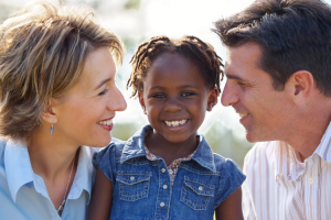 How to Celebrate Adoption Awareness Month blog image. A white woman and man smile and look at their adopted, African American daughter.